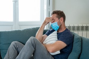 Sad man with protective face mask at home living room couch feeling tired and worried suffering depression amid coronavirus lockdown and social distancing. Mental Health and isolation concept.